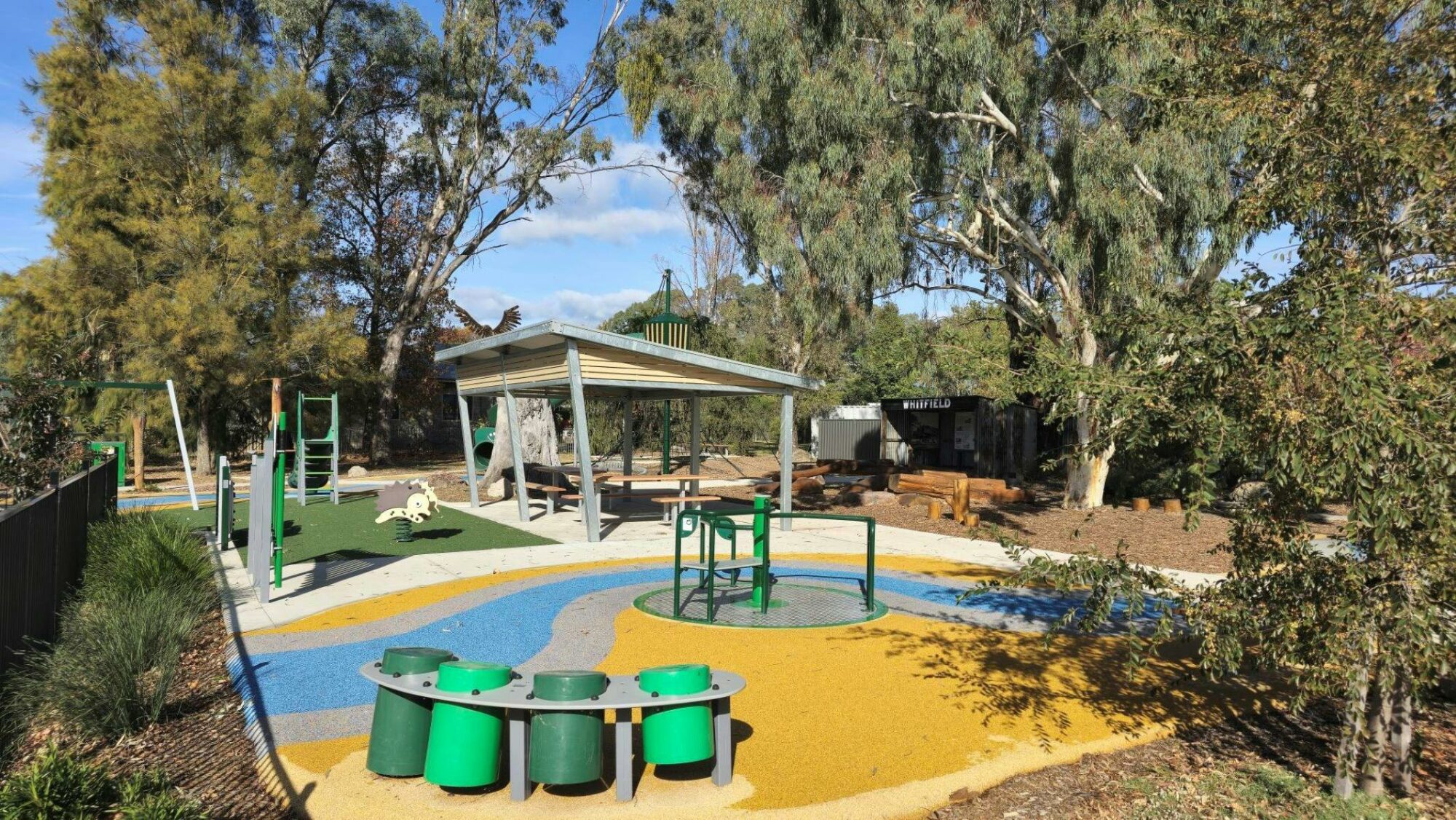 Playground area with green drums on a yellow surface. Shaded seating area in background