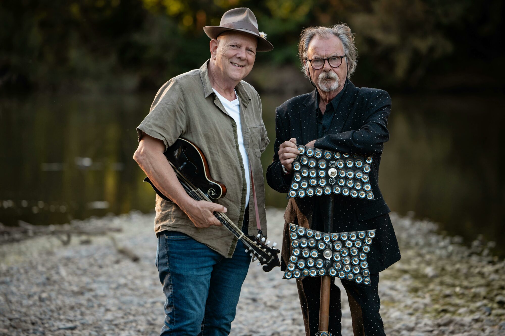 Two men, one holding a mandolin, the other a lagerphone, standing in front of a river.