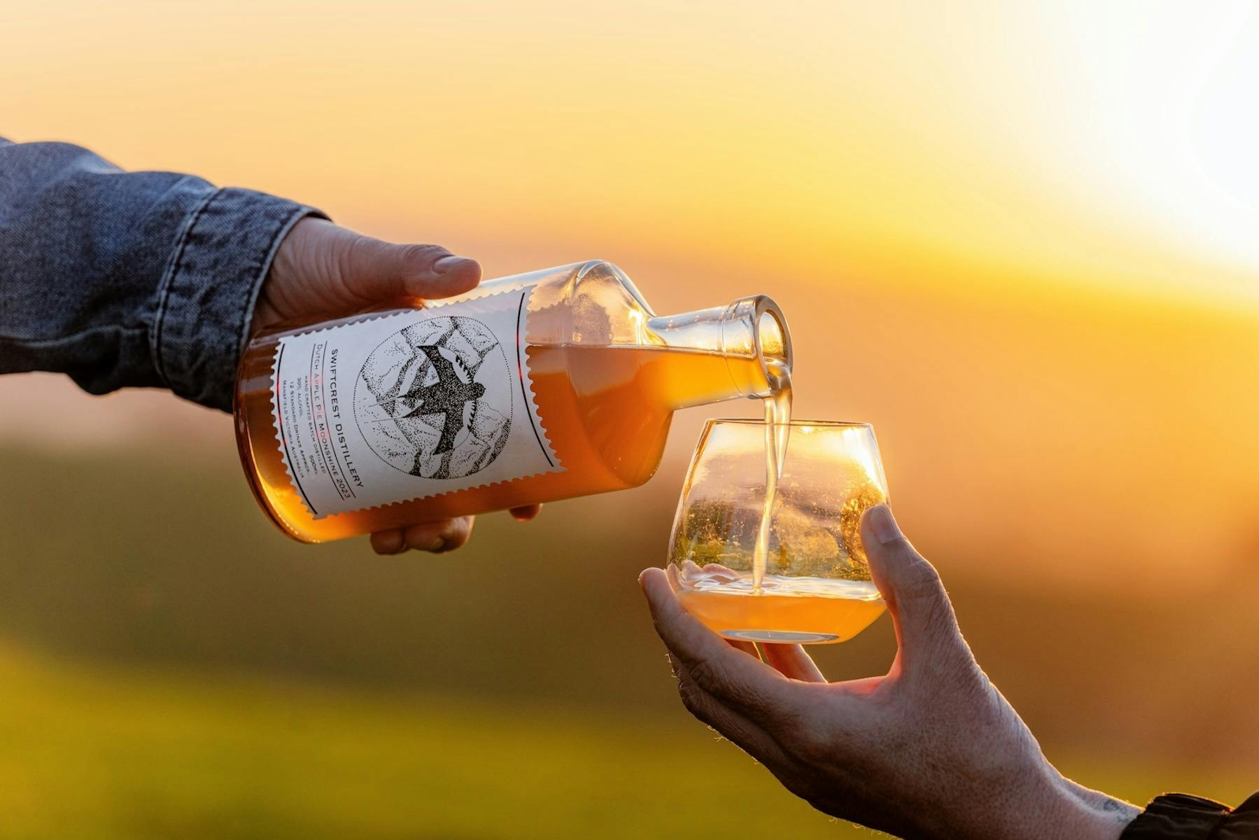 A hand pours amber - coloured spirits from a Swiftcrest Distillery bottle into a glass at sunset.
