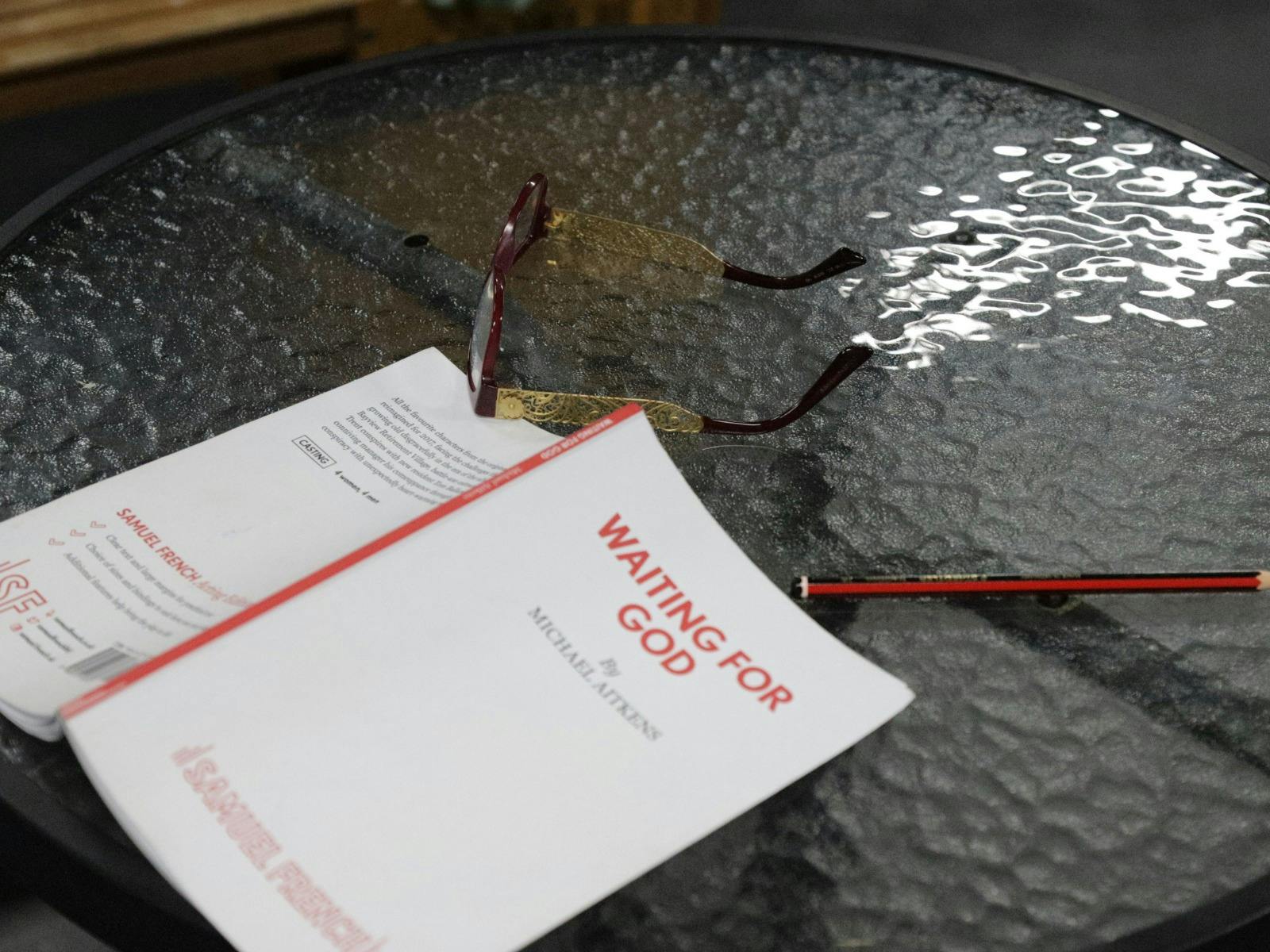 Waiting For God Script facedown on a glass table with a pencil and glasses