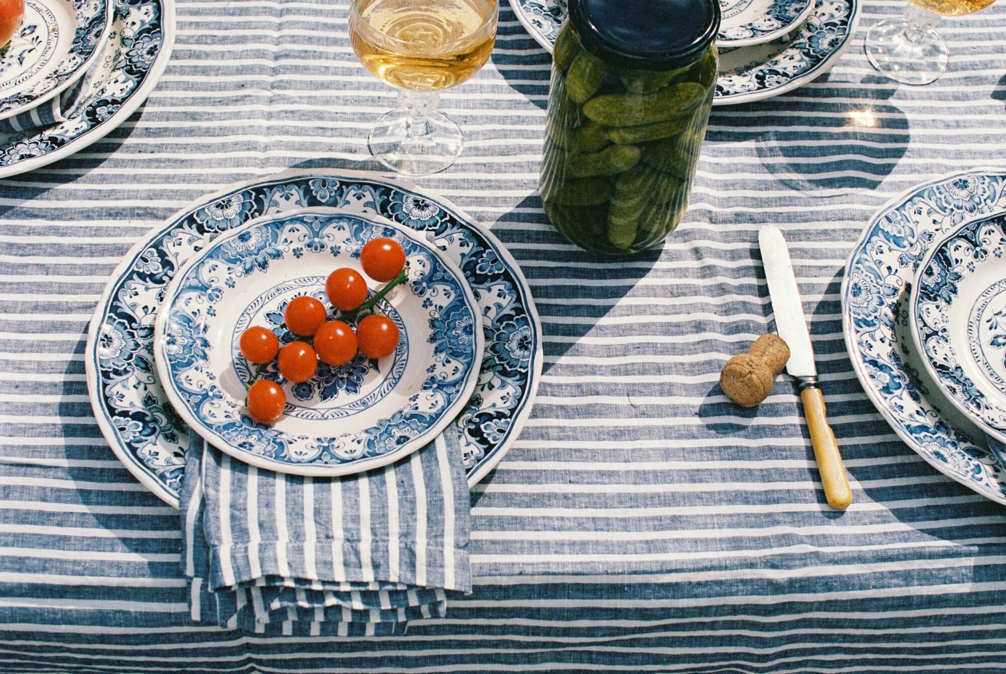 Blue and white table setting with striped linen, cherry tomatoes, pickles, wine glasses, and rustic