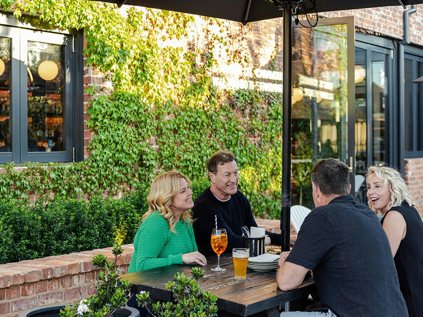 4 people in conversation sitting at a black picnic table, enjoing a beer and an Aperol Spritz