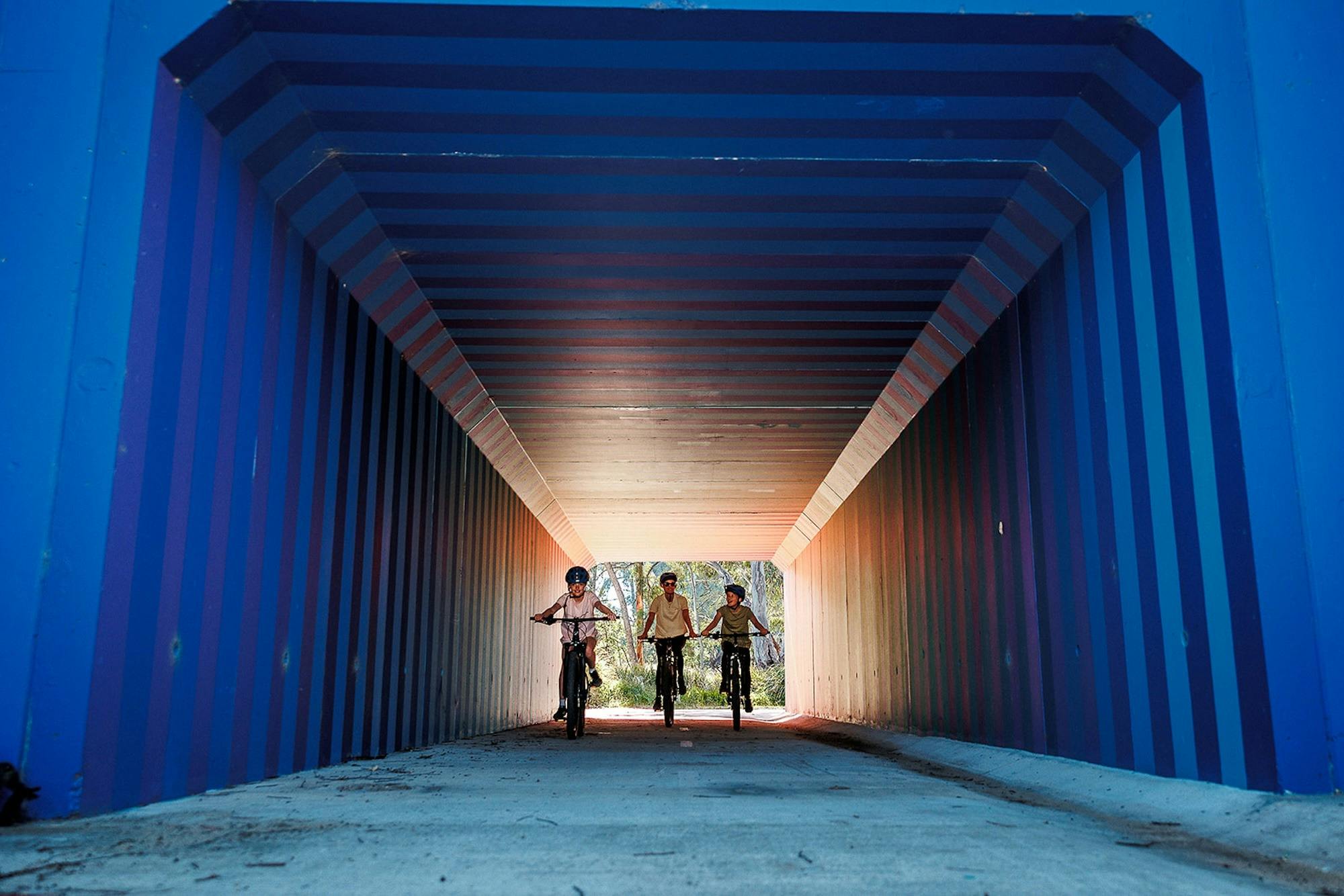 Three cyclists riding through a tunnel painted with blue stripes