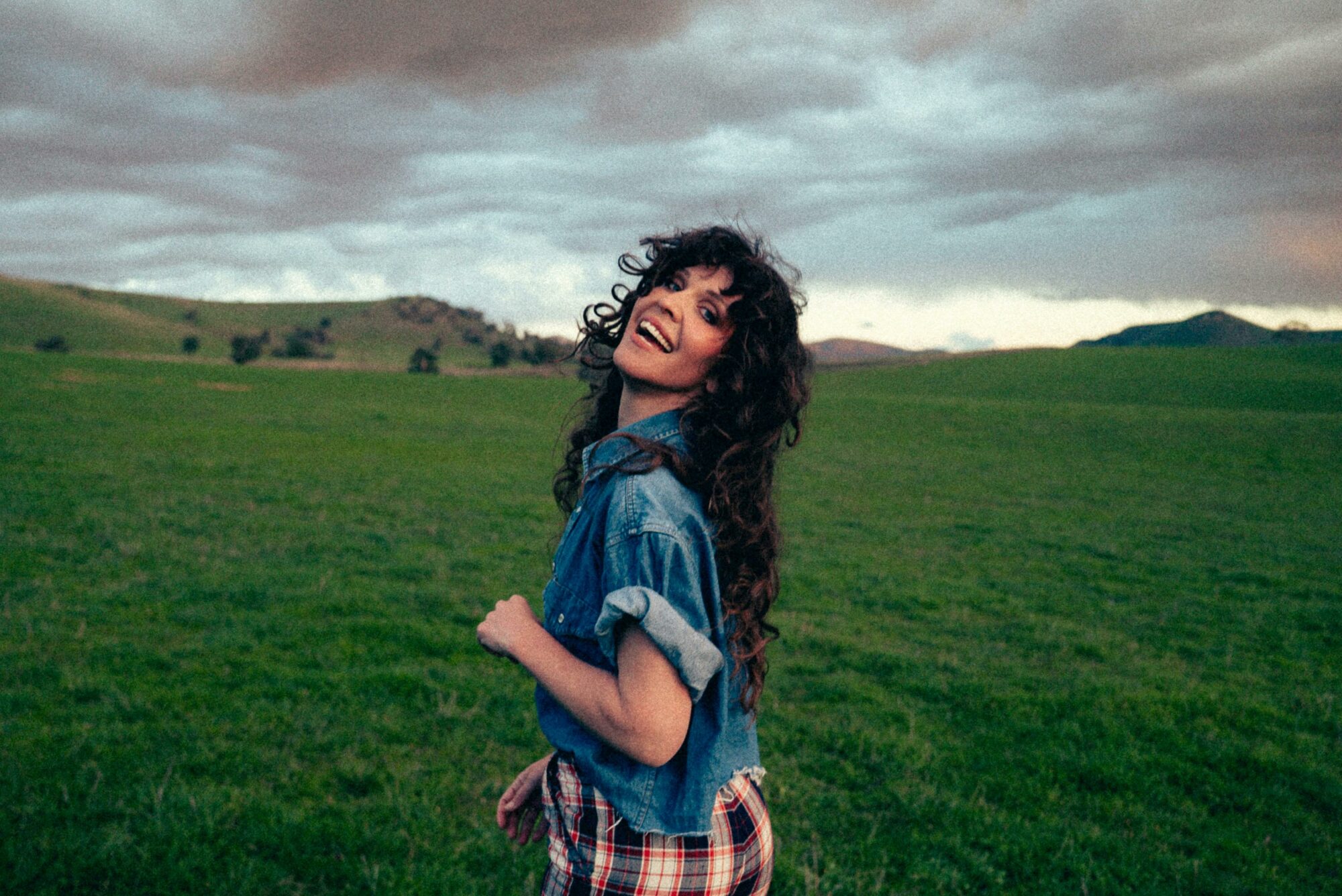 Smiling woman looking over her shoulder, lush green rolling hills & stormy sky in background