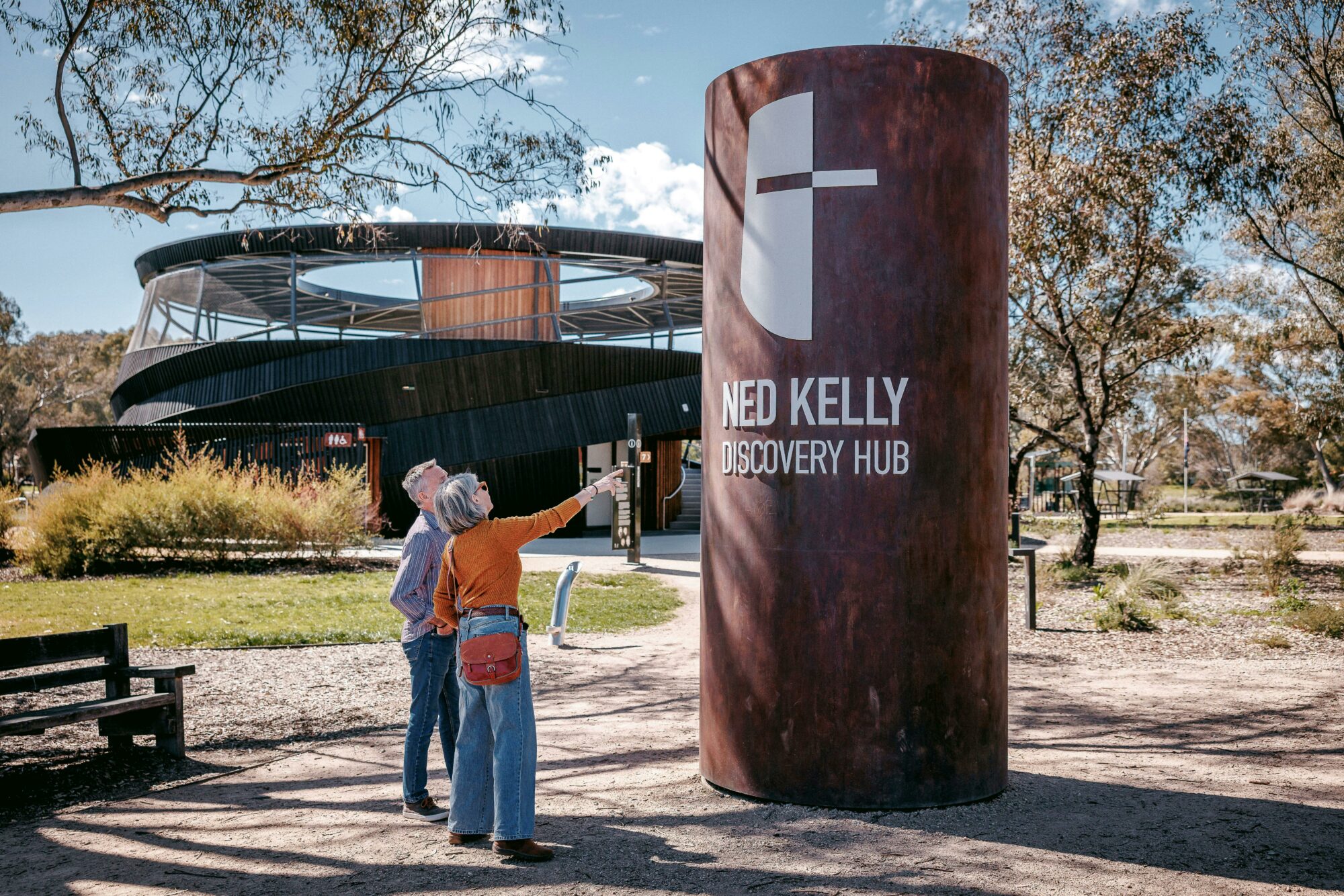 People stand beside a large Ned Kelly Discovery Hub sign, with the modern curved building and tree