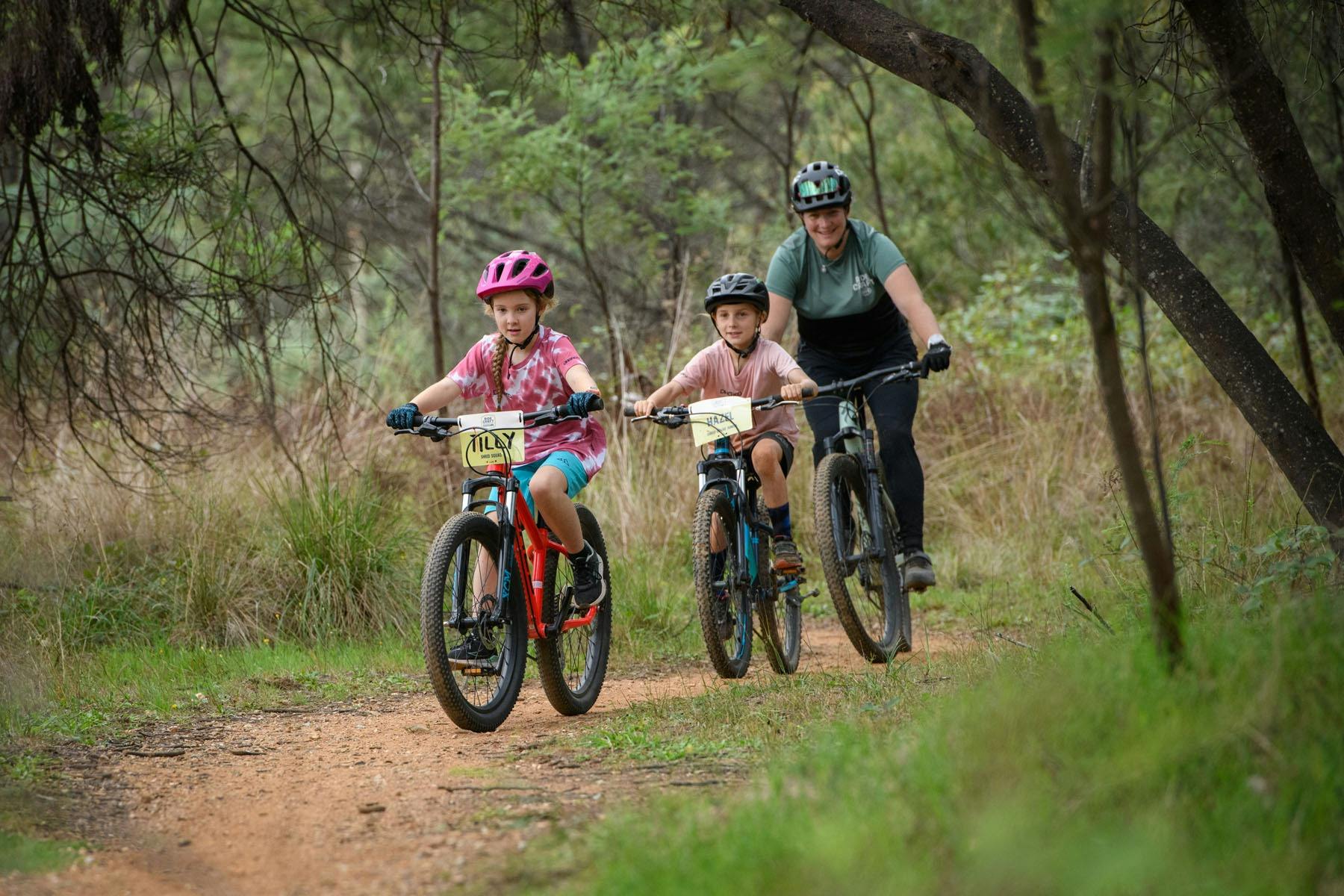 Two young female riders ride down a dirt path, followed by a coach