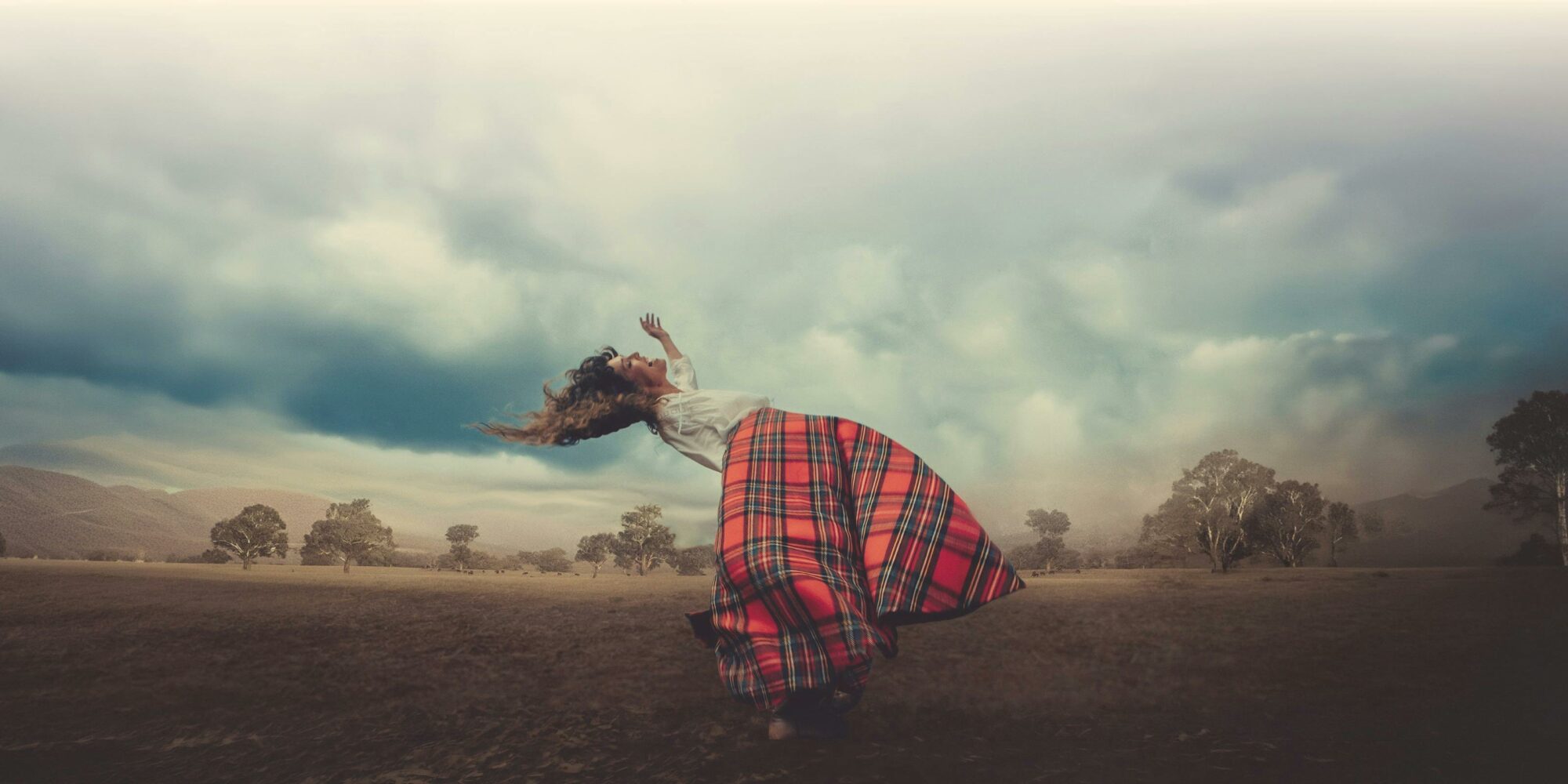 A woman is floating above the ground. She wears a tartan skirt, the sky is stormy & the farmland dry