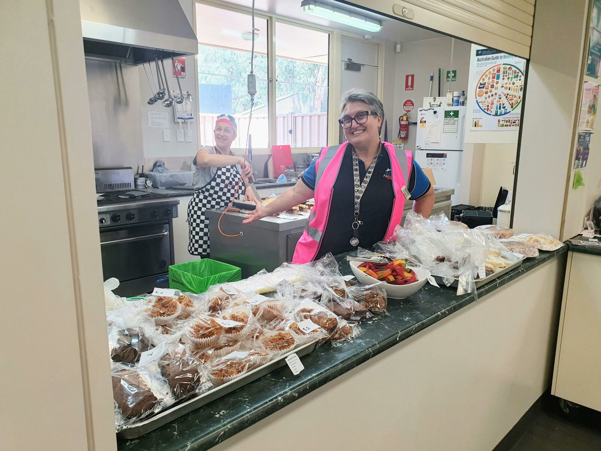 Two smiling people at a bake sale with a range of muffins, cookies and other sweet treats.