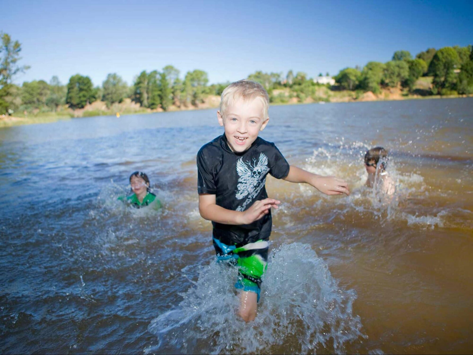 A young blonde boy emerges from a lake smiling. 