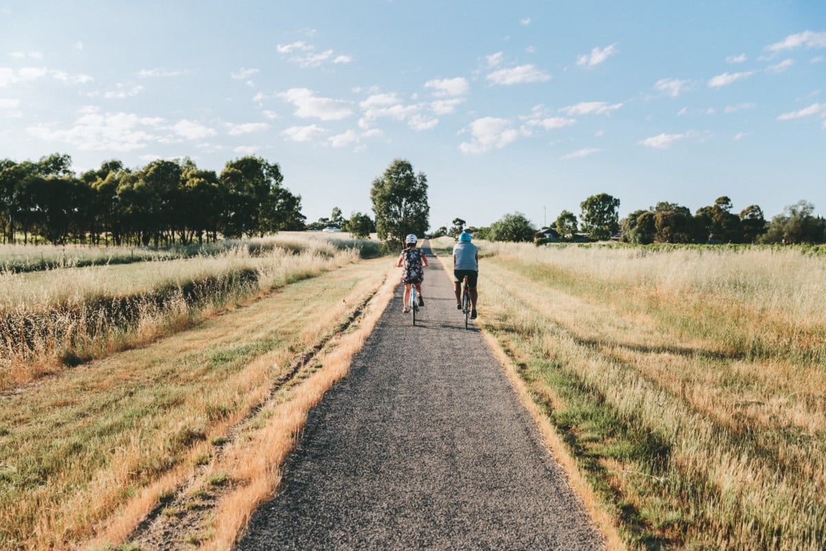 Two people ride bikes along a long, straight trail through a paddock in summer time